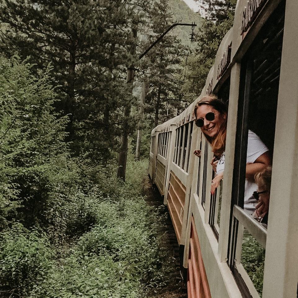ragazza affacciata dal finestrino di un treno che passa in mezzo alle montagne